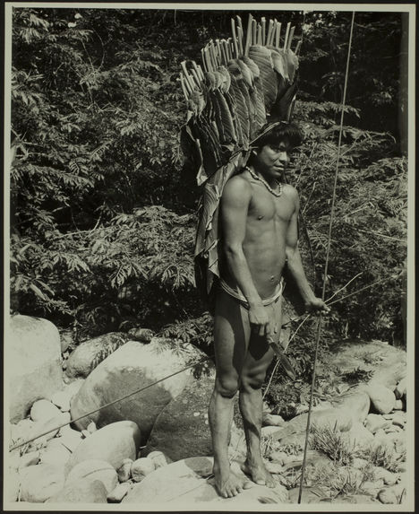 Sans titre [un homme de trois quarts en pied, maintient par la tête et au niveau de la taille un grand panier fait en feuillages et en bois dans le dos. Il tient deux longues tiges en bois pour la pêche ?]